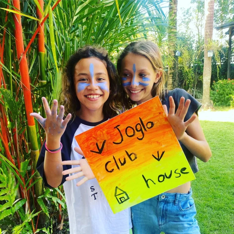Two girls are holding up a bright 'Joglo Clubhouse' hand-painted sign.